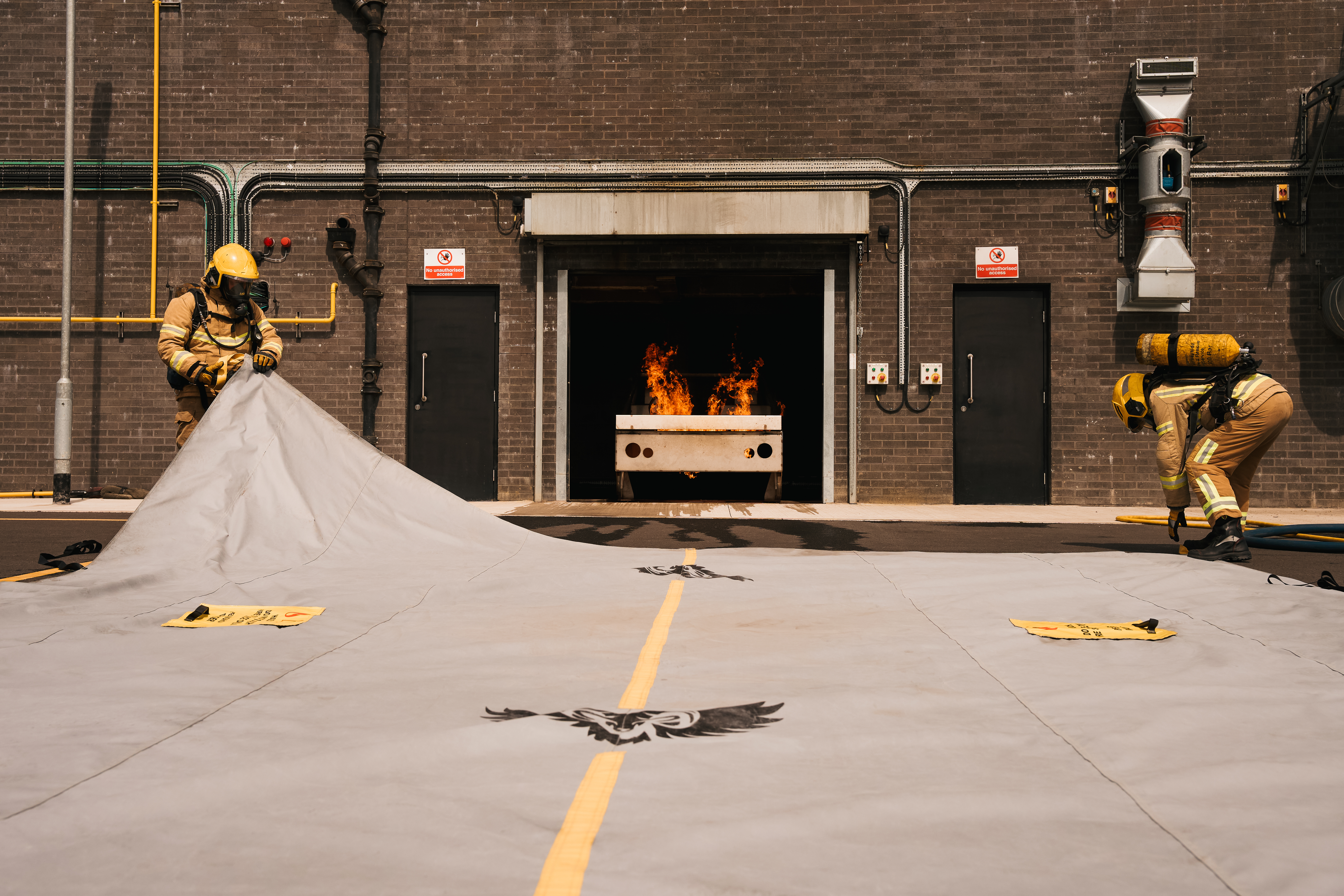 Two firefighters in full PPE preparing the Golden Fleece EV containment blanket in front of an active flame at a loading bay — real deployment of the EV thermal-runaway containment system, ram-and-wings brand mark and yellow alignment stripe visible on the spread blanket.